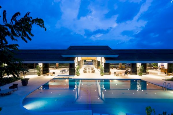 A luxurious modern building with large glass windows is in front of a beautifully lit swimming pool at dusk.