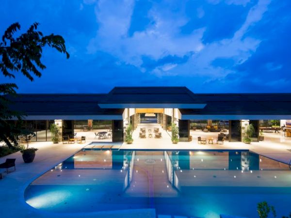 A luxurious modern building with large glass windows is in front of a beautifully lit swimming pool at dusk.
