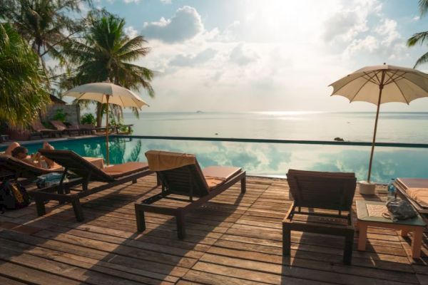 A serene poolside setting with deck chairs, umbrellas, palm trees, and a calm sea under a bright sky, inviting relaxation.