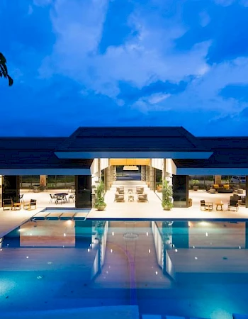 An illuminated modern building with a large swimming pool at dusk reflects the deep blue sky and surrounding lights.