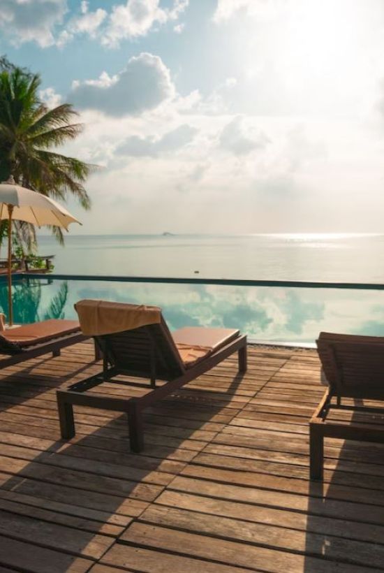 A serene poolside scene with sun loungers, umbrellas, palm trees, and a tranquil ocean view under a bright sky on a wooden deck.