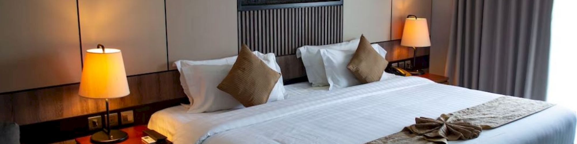 A cozy hotel room with a large bed, two bedside tables with lamps, a wall-mounted decor piece, and curtains on the window.