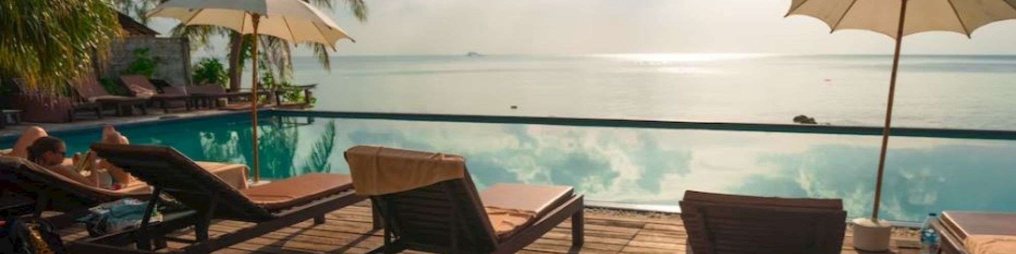 A serene poolside scene with lounge chairs, umbrellas, and palm trees overlooking a calm sea under a clear, sunny sky.