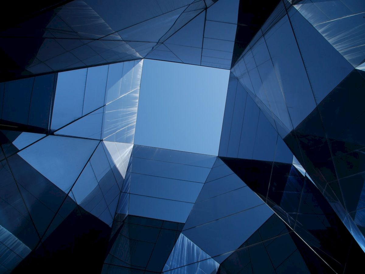 The image shows an upward view through a geometric structure with reflective glass panels, forming a pattern against a clear blue sky.