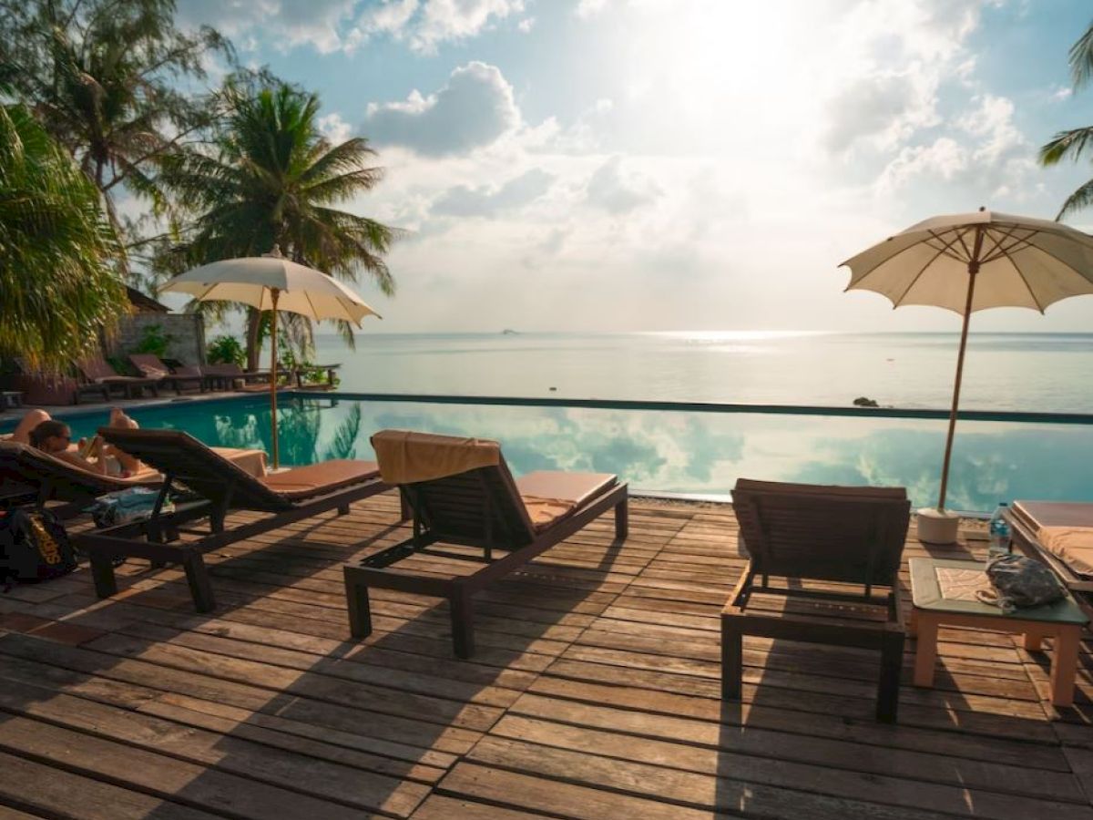 A serene poolside scene with lounge chairs, umbrellas, and palm trees overlooking a calm body of water under a bright sky.