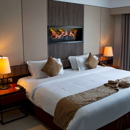 A hotel room with a large bed, two side lamps, an artwork above the bed, and a window with curtains on the right side of the room.