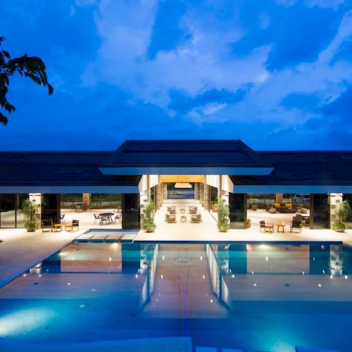 A luxurious, illuminated pool and modern building surrounded by trees under a blue evening sky, reflecting a tranquil ambiance.