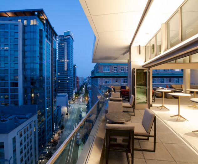 A modern rooftop terrace with seating, overlooking a city street flanked by tall buildings during the evening.