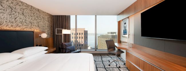 A modern hotel room with a large bed, desk, chair, large TV, and a view of the city skyline through wide windows ending the sentence.