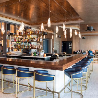 A modern bar with blue stools, a fully stocked shelf, and hanging lights. There's seating and tasteful decor in the spacious lounge area.