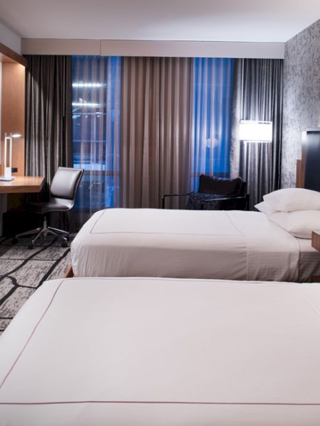 The image shows a modern hotel room with two beds, a wall-mounted TV, desk, chair, and stylish decor, including patterned walls and curtains.