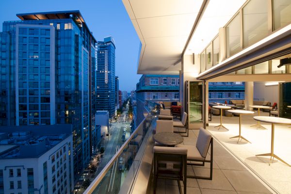 A modern cityscape at dusk with tall buildings and a sleek, open terrace featuring tables and chairs along the railing.