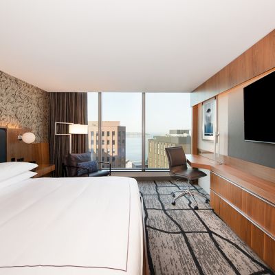 A modern hotel room with a large bed, wall-mounted TV, desk, and city view through a window, decorated in neutral tones.