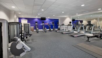 The image shows a gym with treadmills, stationary bikes, and weight machines in a spacious room with mirrors and a motivational wall sign.