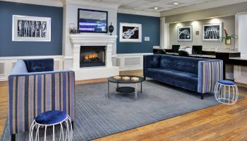 The image shows a modern lobby with blue sofas, a fireplace, framed art, and a reception desk in the background.