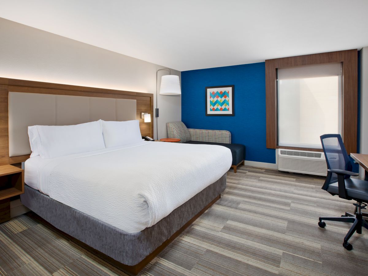 A modern hotel room with a large bed, desk, chair, sofa, and wall art. Neutral tones and a blue accent wall enhance the space.