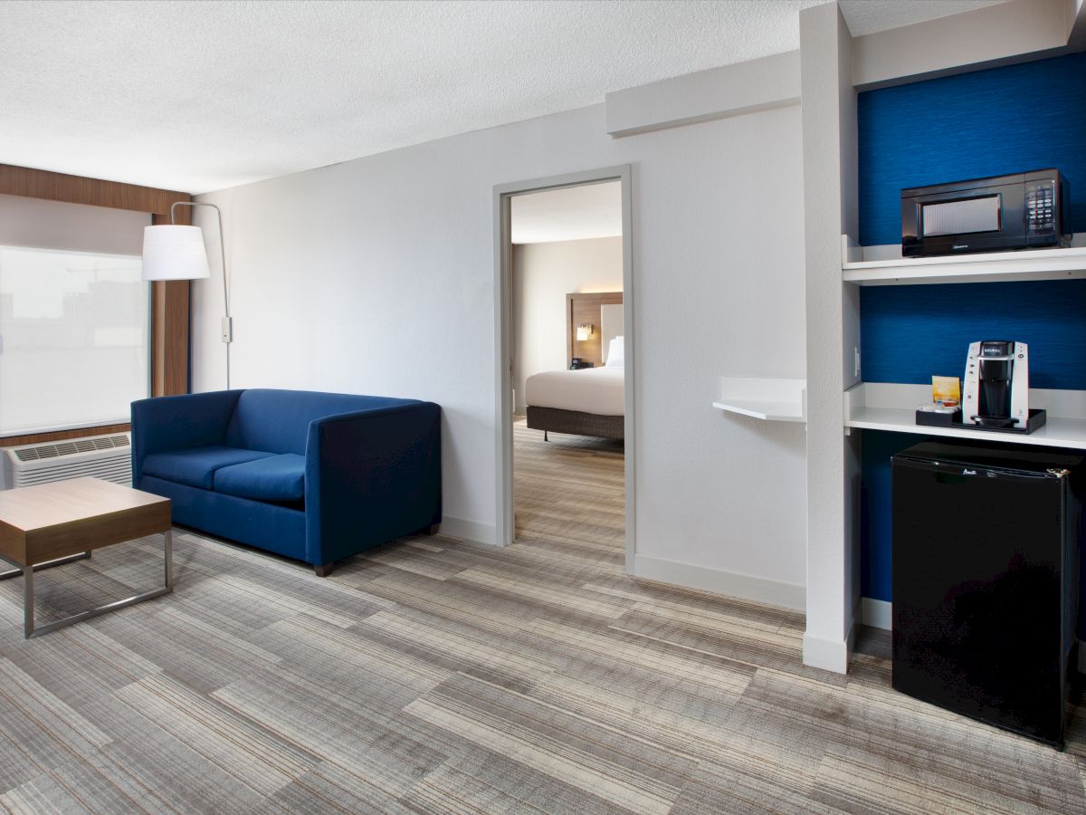 The image shows a modern hotel room with a blue sofa, a small coffee table, a mini-fridge, a microwave, and a coffee maker on a shelf.