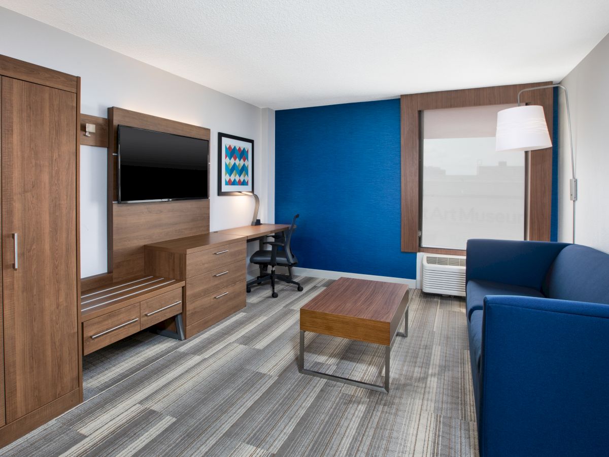 A modern room with wooden furniture, a wall-mounted TV, desk, chair, blue accents, sofa, and window with a city view.
