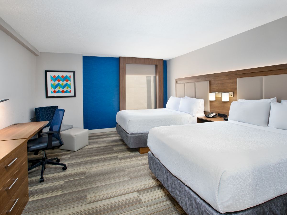 The image shows a hotel room with two large beds, a desk with a chair, a TV, a piece of artwork on the wall, and a wood-textured floor.