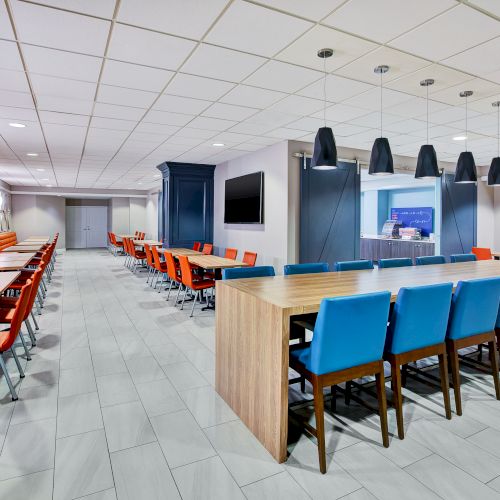 The image shows a modern dining area with red and blue chairs, wooden tables, pendant lights, and tiled floors, creating an inviting space.