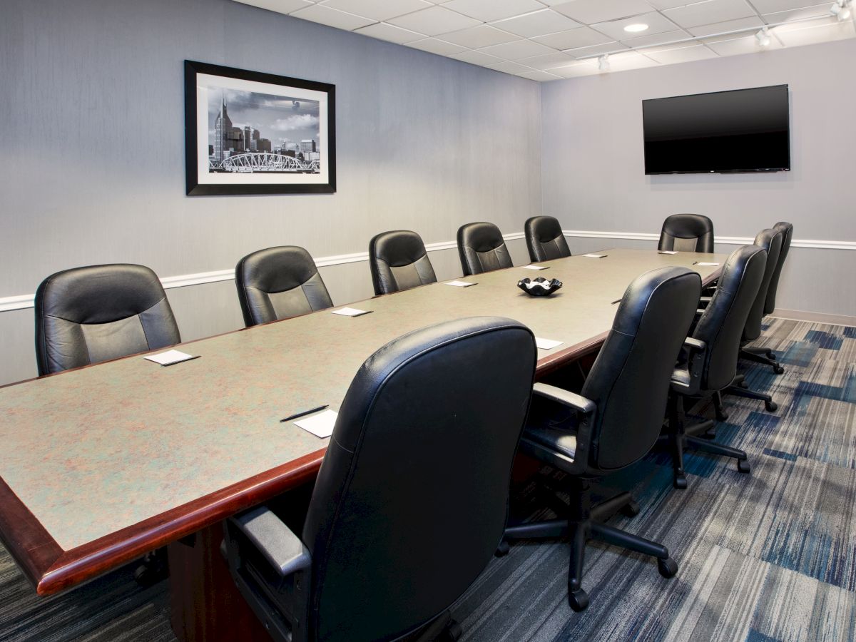 A modern conference room with a long table, black chairs, wall art, and a TV screen on the wall, featuring a patterned carpet floor.