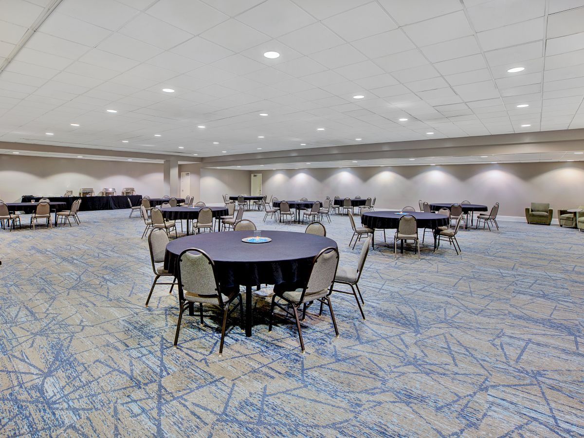 The image shows a spacious conference room with round tables, chairs, and a patterned carpet, suggesting a setup for an event or meeting.