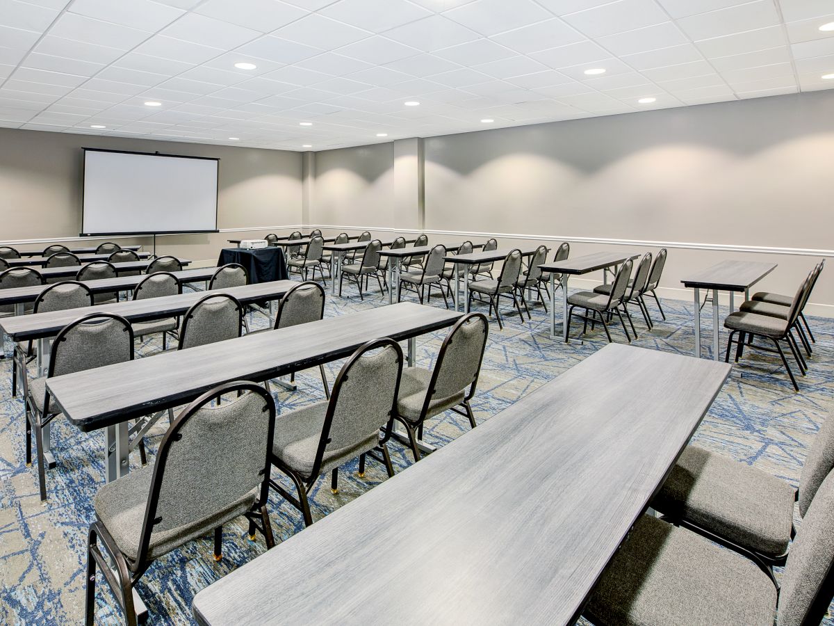 The image shows a conference room setup with rows of tables and chairs facing a projection screen, ideal for meetings or presentations.