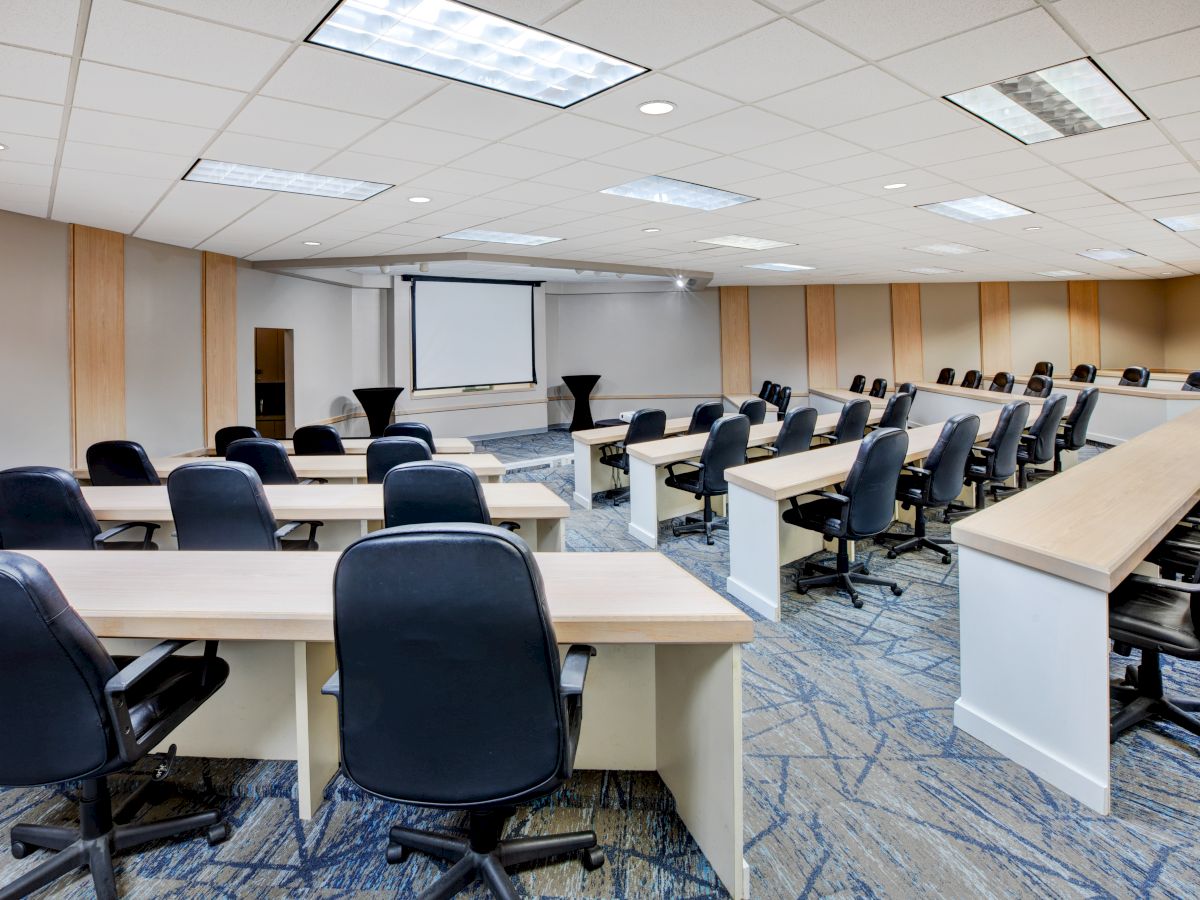 A modern conference room with rows of tables and chairs, a projector screen, and bright overhead lighting.