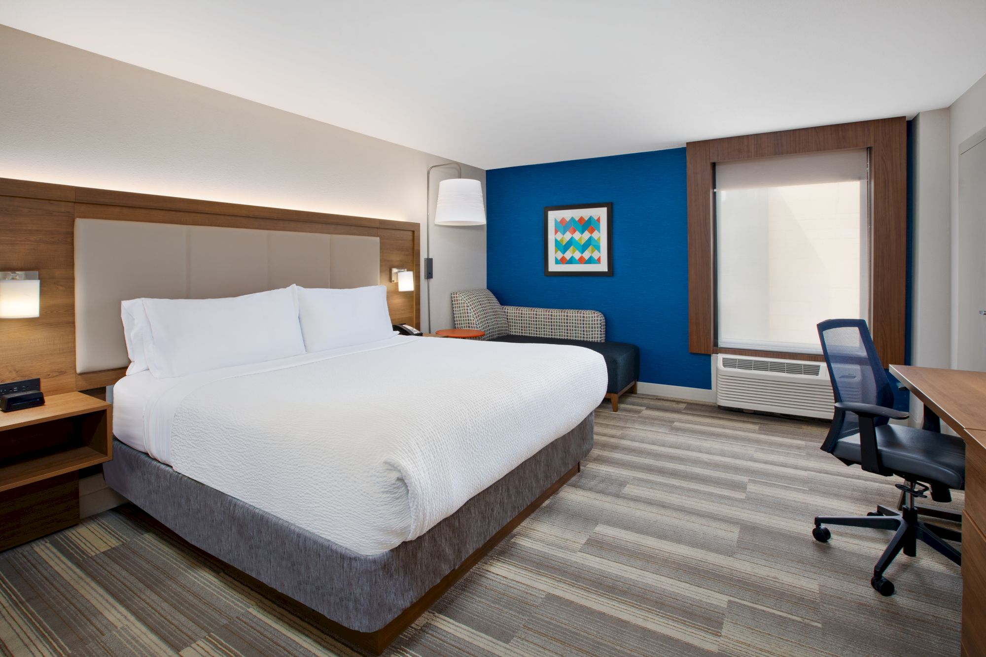 A modern hotel room with a king-size bed, a blue accent wall, a chair, desk with a chair, and a window with a shade drawn.