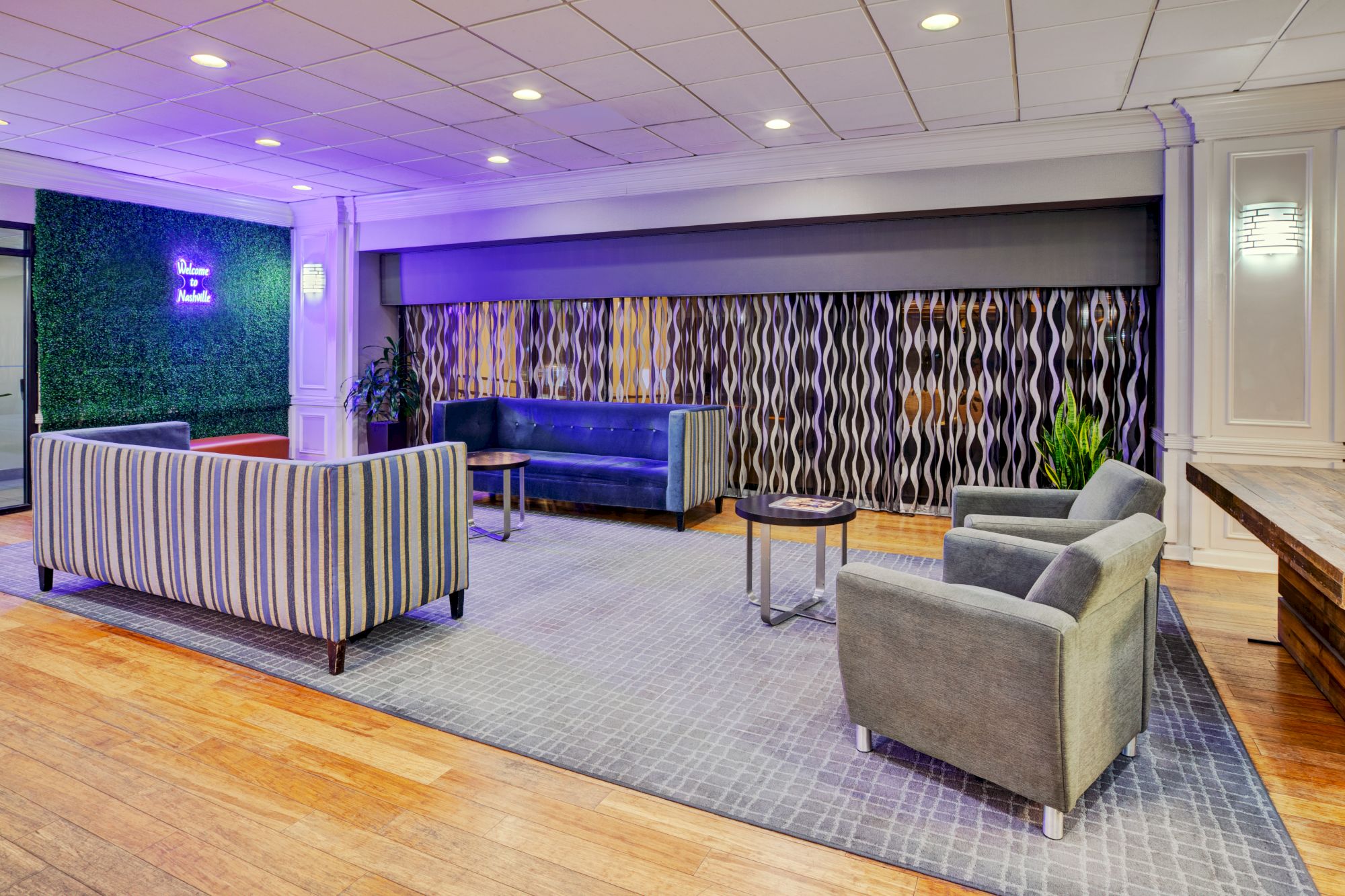 A modern lounge area features striped and gray seating, accent tables, a patterned curtain, and a wall with a neon sign on greenery.