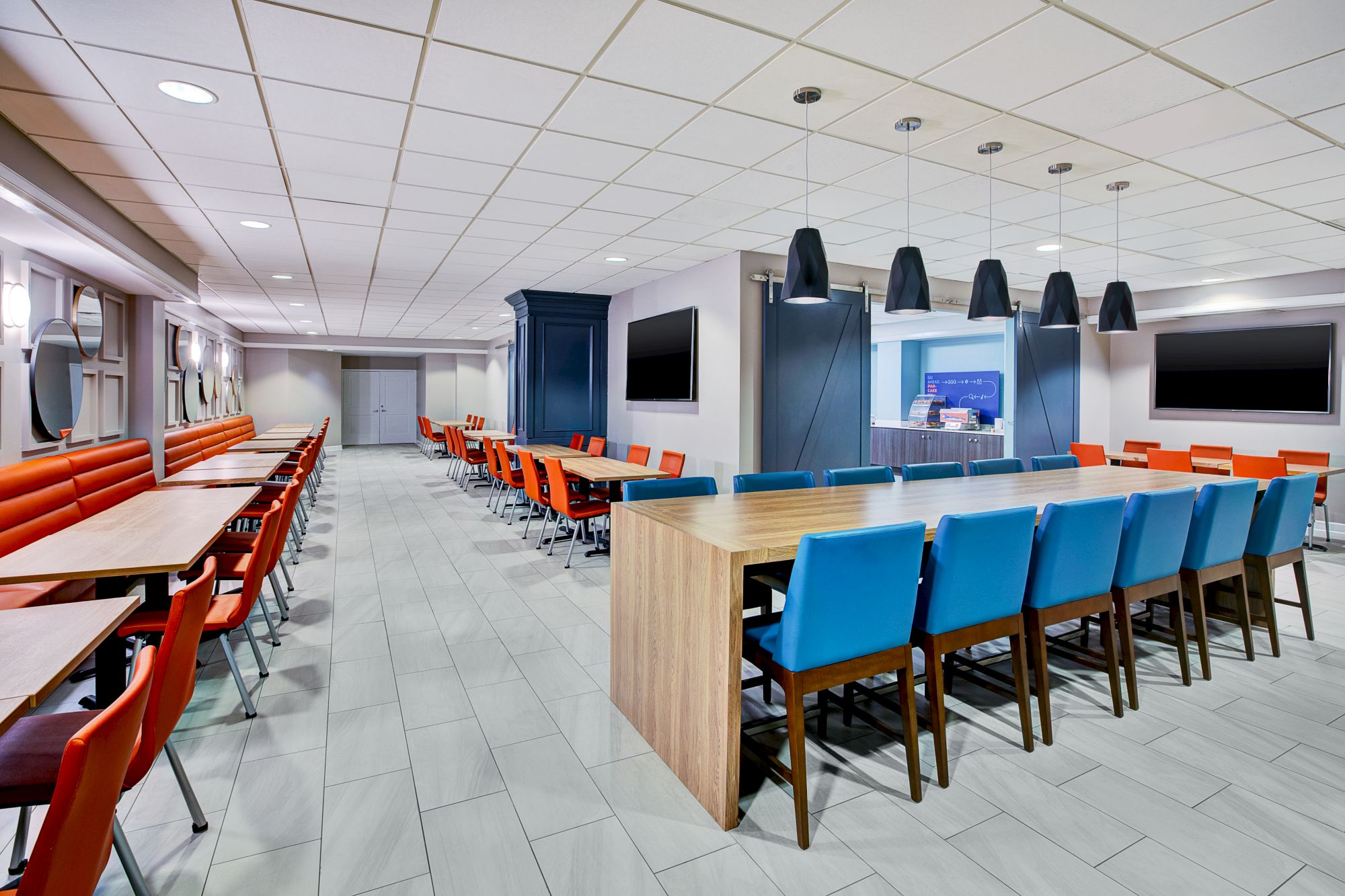 The image shows a modern dining area with orange and blue chairs, long tables, overhead lighting, and a tiled floor.