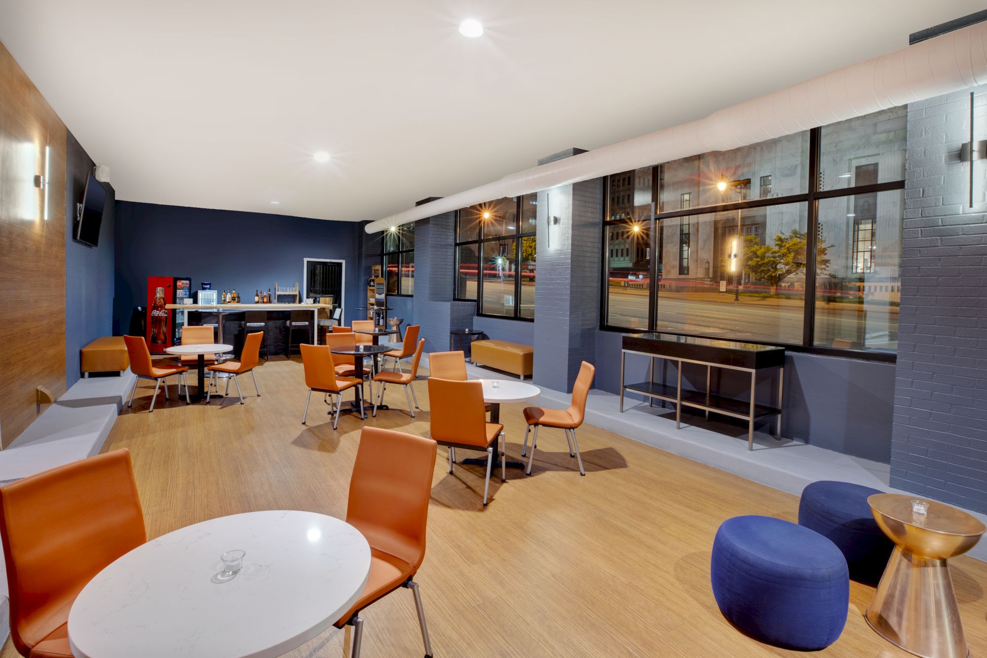 A modern lounge with orange chairs, round tables, and large windows showing an urban night scene, featuring a TV and vending machine.