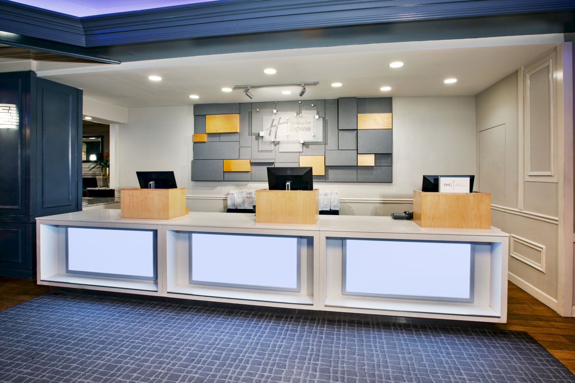 A modern hotel reception desk with three workstations, a geometric art piece on the wall, and a softly lit ambient environment.