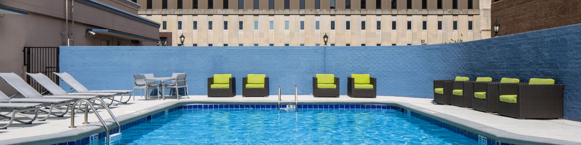 A rooftop pool with blue water, surrounded by lounge chairs and green-cushioned seating, set against a blue wall and urban backdrop.