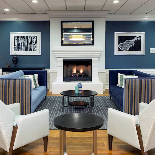 A modern lounge with blue sofas, white armchairs, a fireplace, and framed art on the wall, creating a cozy atmosphere.