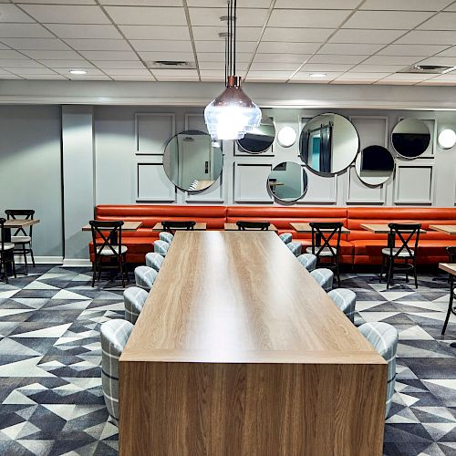 A modern dining area with a long wooden table, chairs, geometric carpet, orange booth seating, and circular mirrors on the wall.