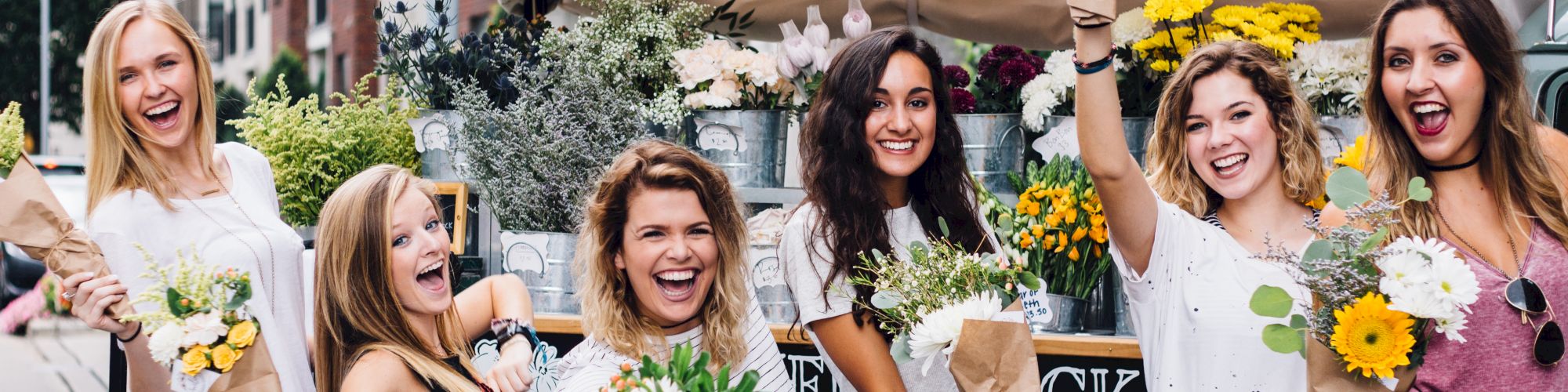 A group of six smiling women holding bouquets, posing in front of a flower stall on a city street, enjoying a sunny day.