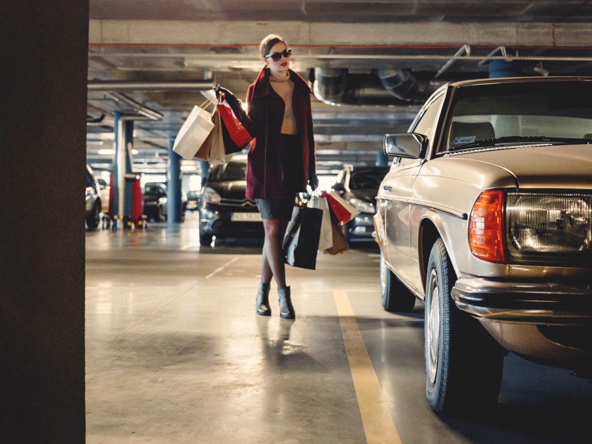 A person in a parking garage is carrying several shopping bags, walking past a parked car with vintage styling.