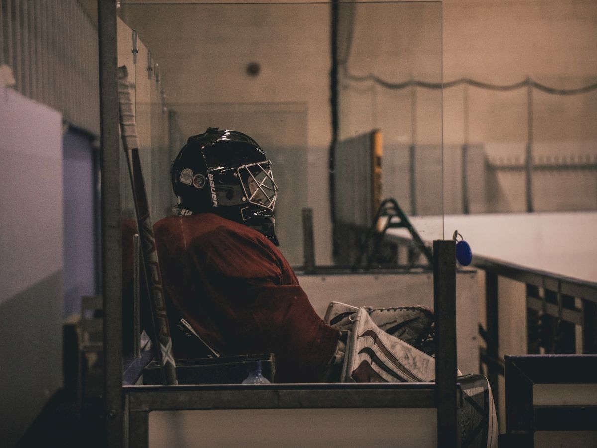 A hockey goalie in gear sits alone on a bench in a rink, surrounded by glass barriers.