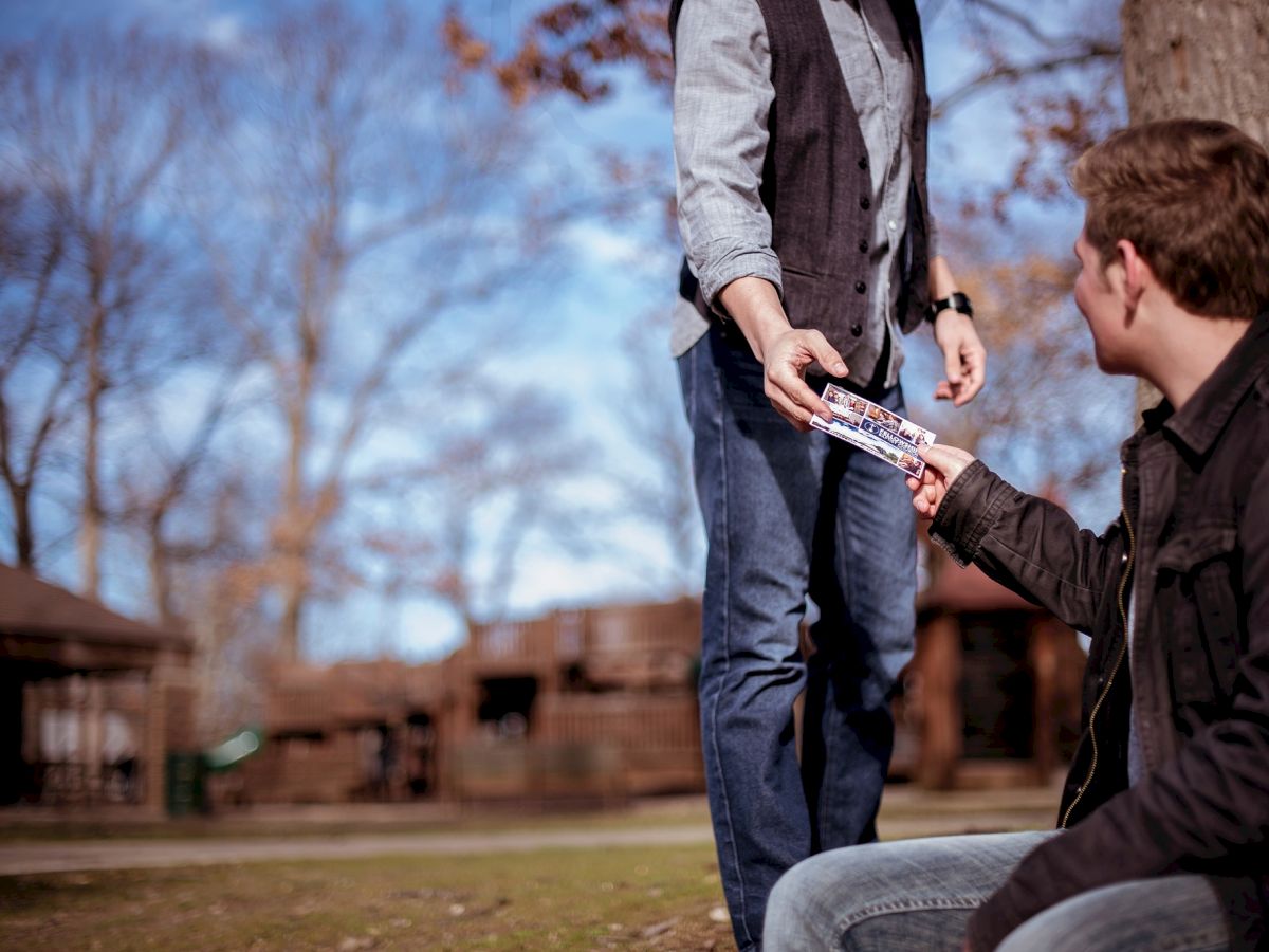 One person hands a snack bar to another, outdoors with trees and blue sky in the background.