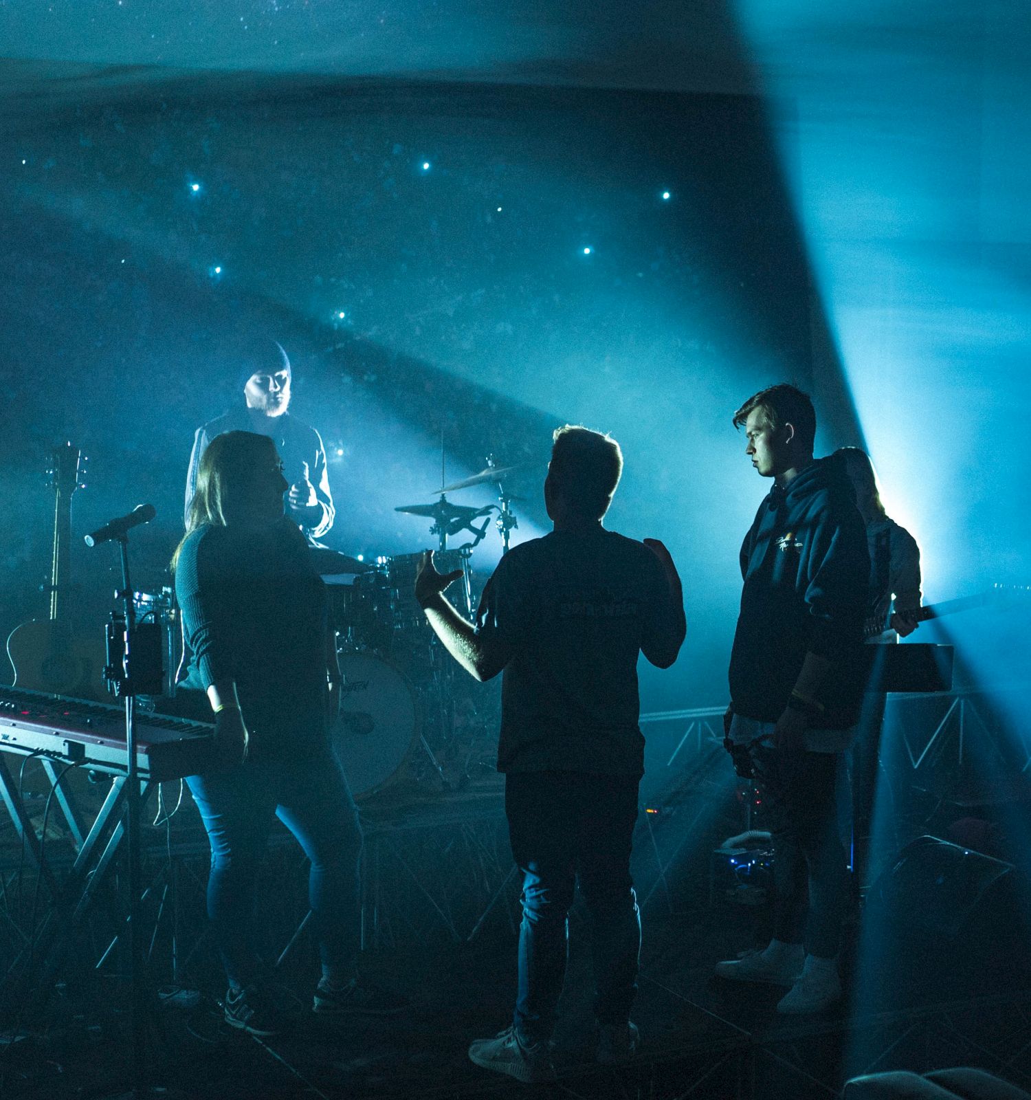 A band rehearses in a dimly lit room with instruments and blue lighting, creating a moody atmosphere on stage.