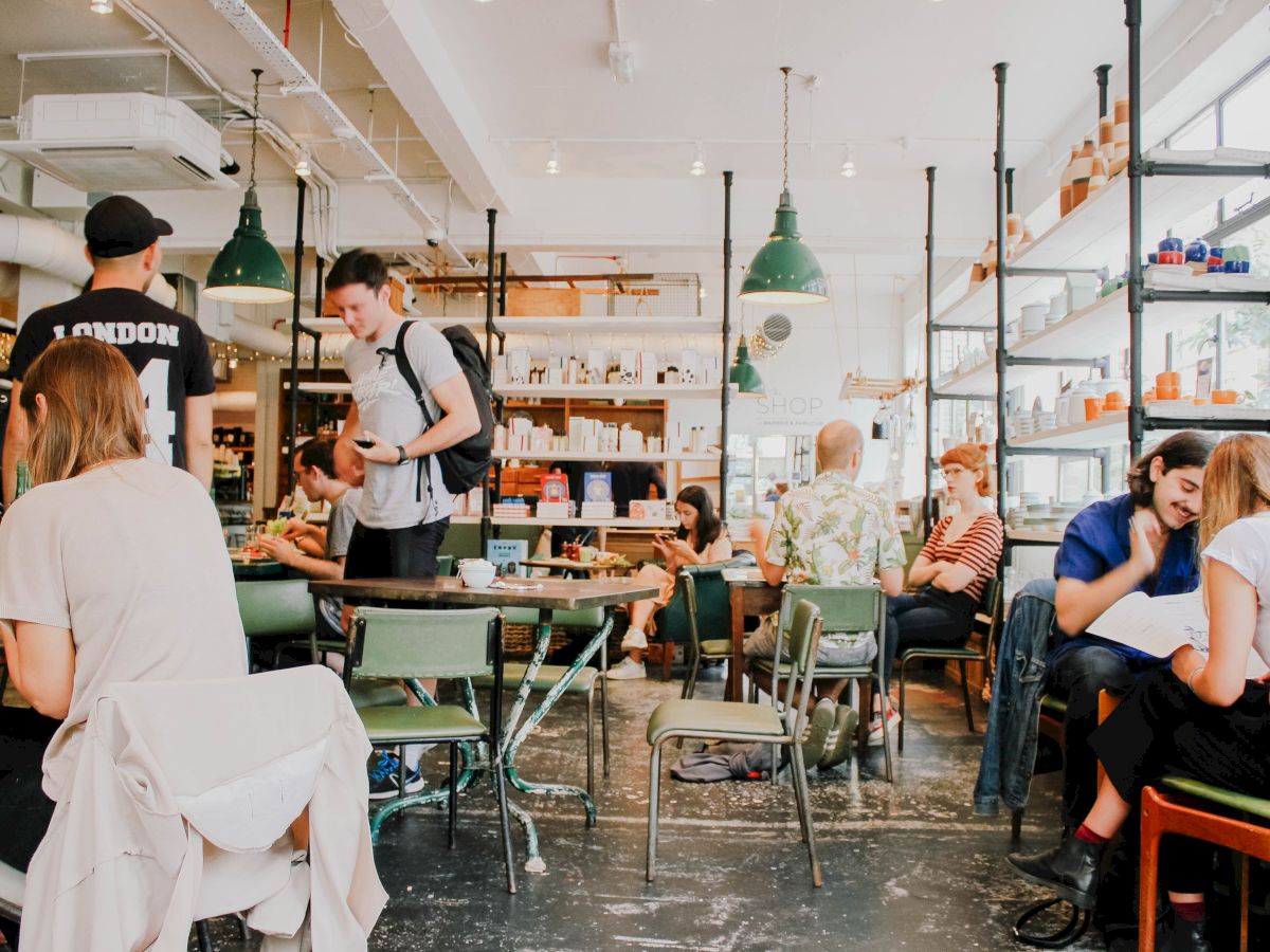 The image shows a busy café with people sitting and chatting at tables, surrounded by shelves and hanging lights.