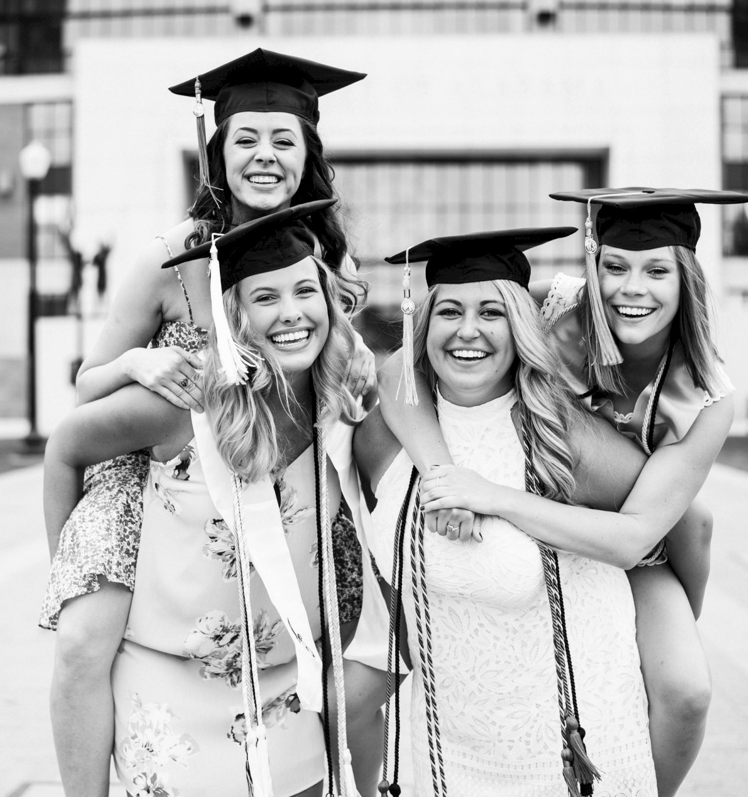 Graduates celebrate with piggyback rides, wearing caps and gowns, smiling joyfully in front of a large building.