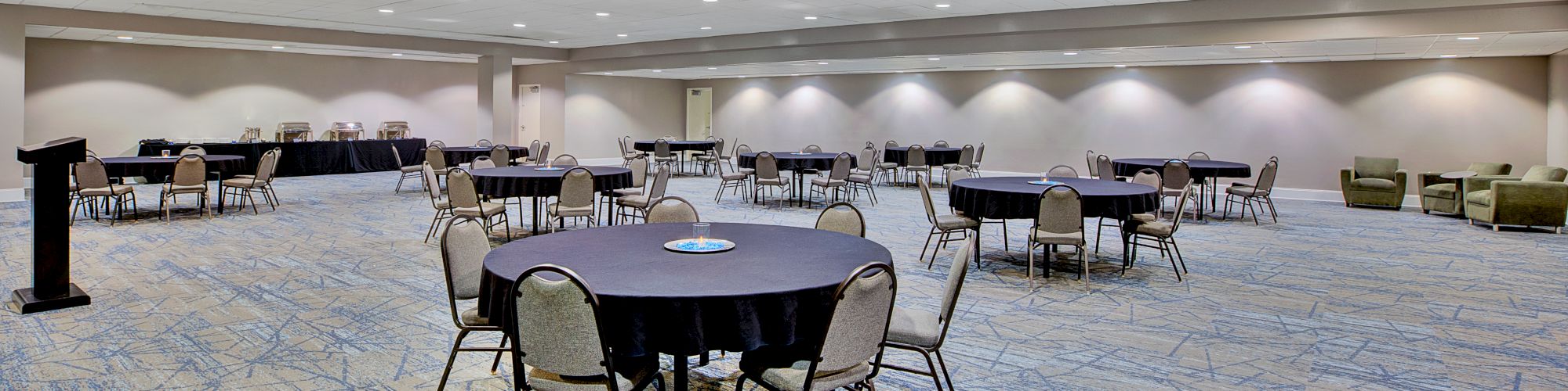 A large empty banquet hall with round tables, chairs, and patterned carpet, under bright ceiling lights.
