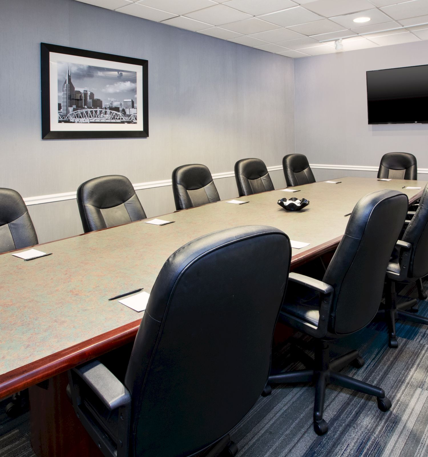 A modern conference room with a long table, surrounded by black chairs, a wall-mounted TV, and a framed picture on the wall.