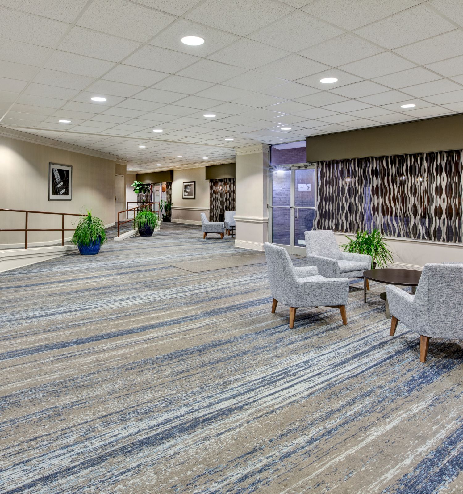 The image shows a modern lobby with patterned carpet, chairs around tables, potted plants, and decorative elements lining the walls.