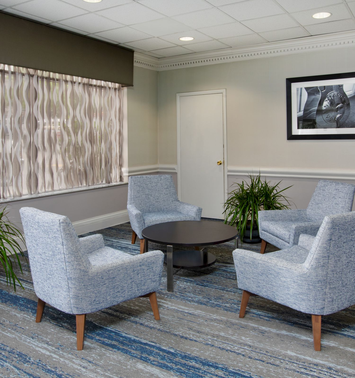 A waiting area with four gray chairs around a circular table, two plants, a window with wavy curtains, and a black-and-white photo on the wall.