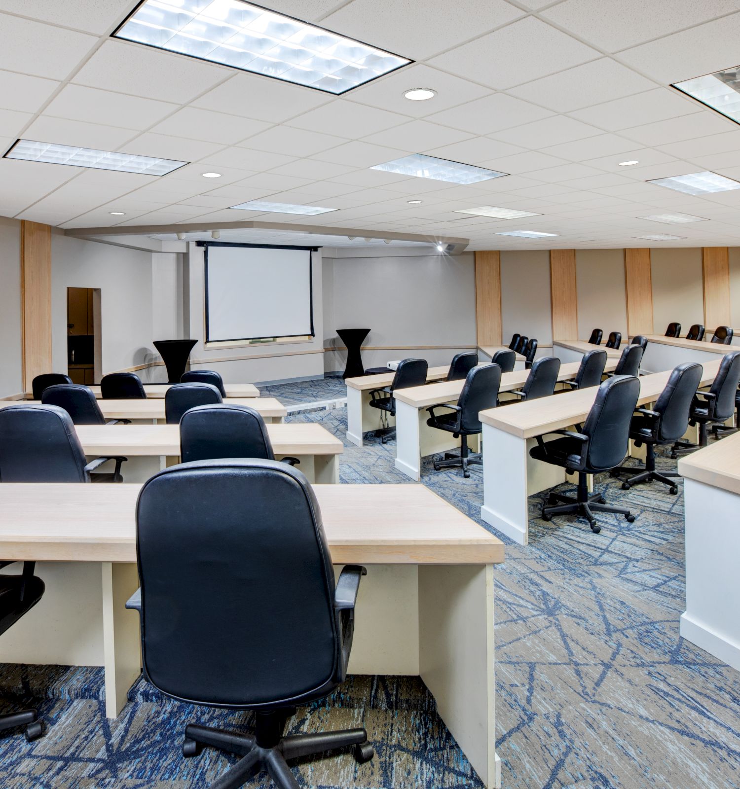 The image shows a conference room setup with rows of tables and chairs facing a screen at the front of the room.