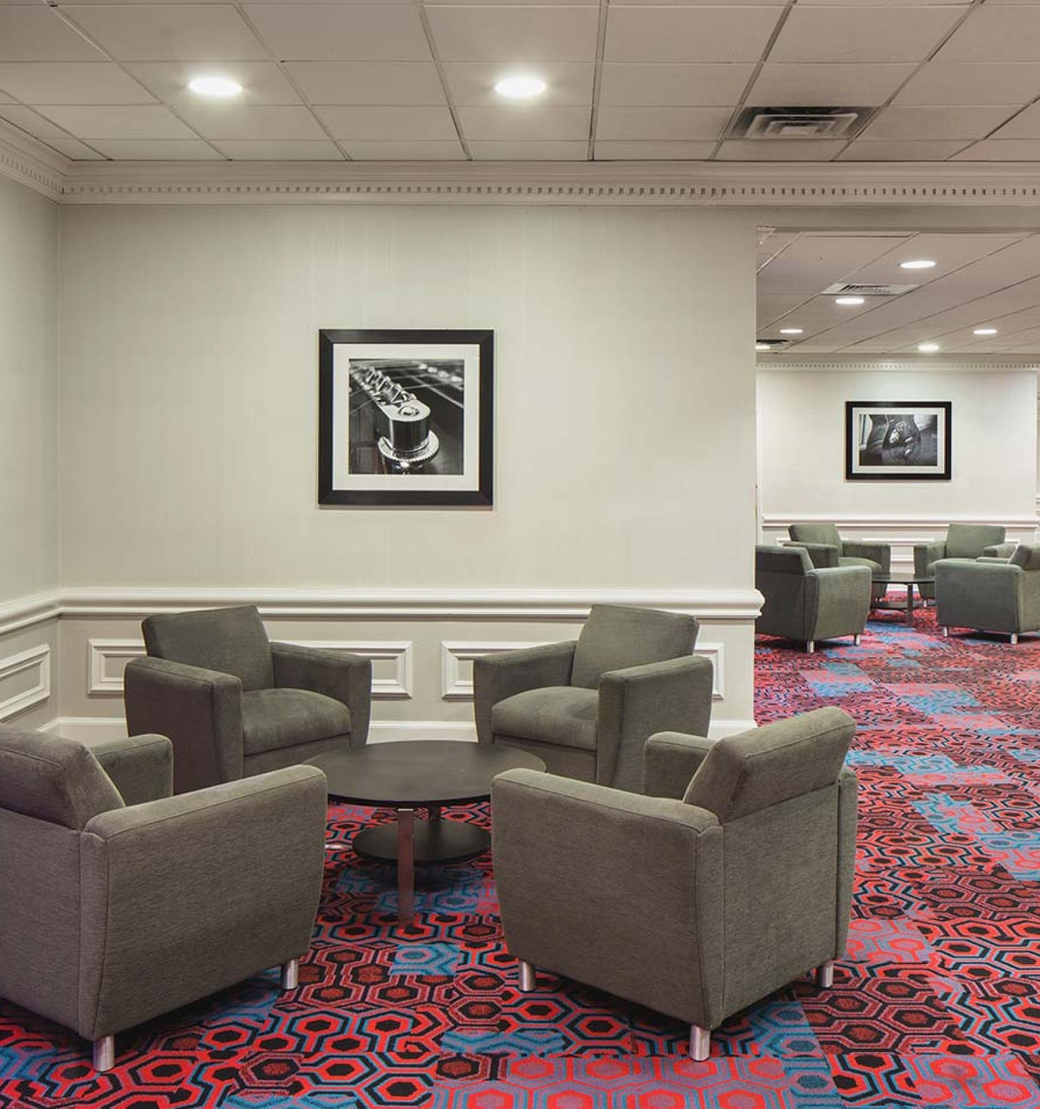 The image shows a lounge area with gray armchairs, a round table, colorful carpet, framed artwork, and curtain-covered windows.