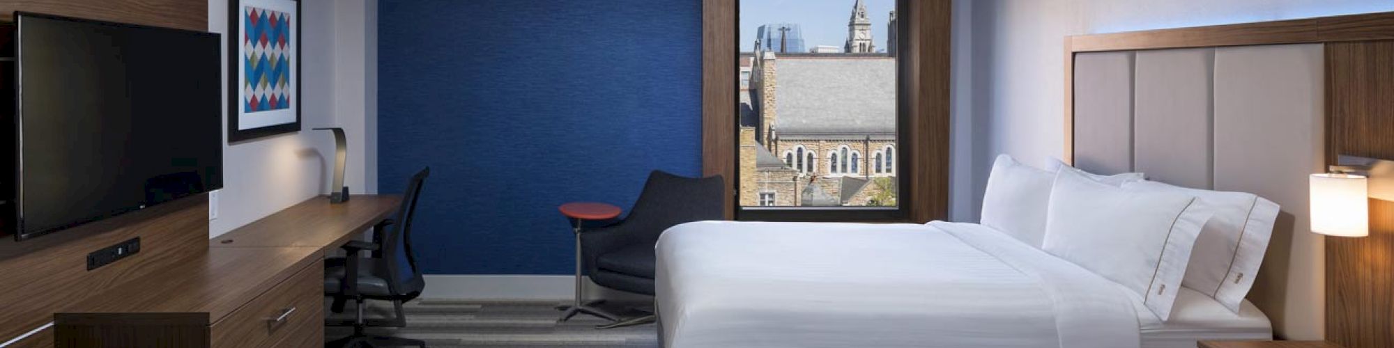 The image shows a hotel room with two beds, a desk, chair, TV, and a window with a view. The room has modern decor and carpeted floor.